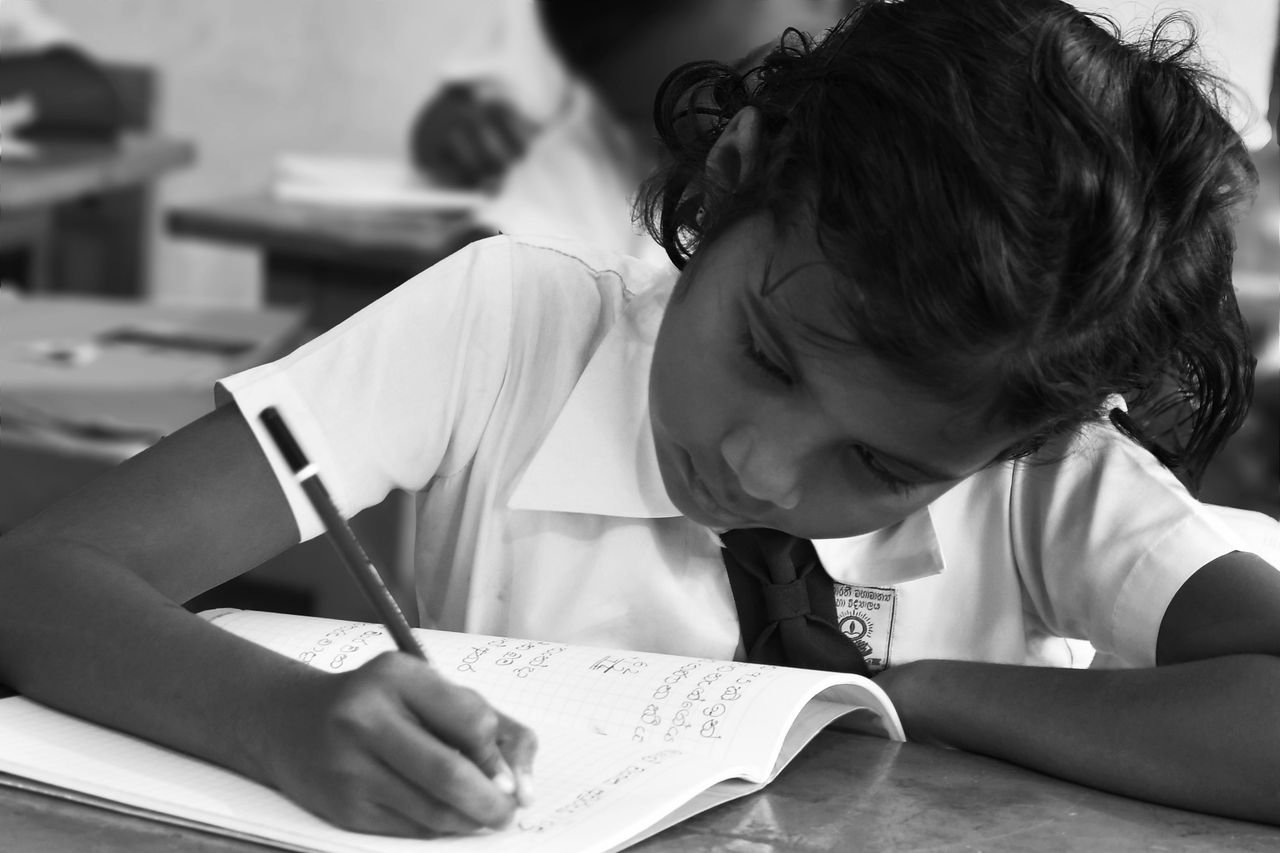 Black and white image of a young girl writing diligently in a classroom setting.